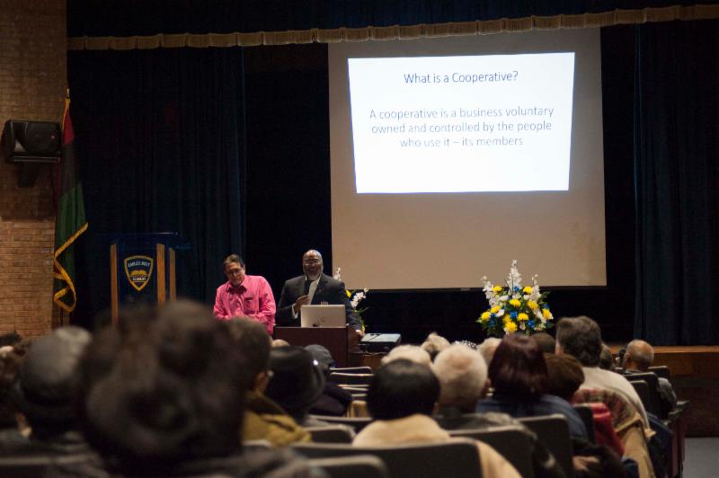 Photo of speakers presenting to an auditorium, showing a slide defining cooperatives